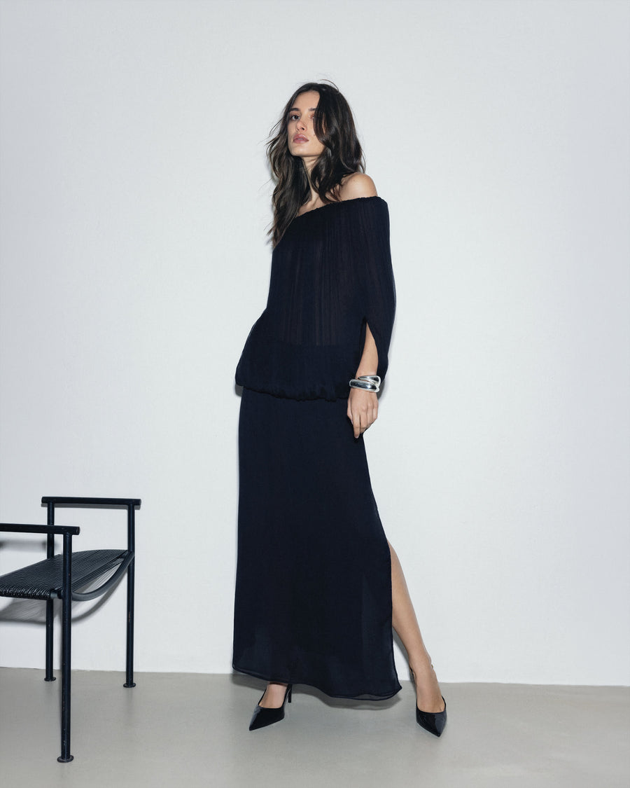 Woman wearing a black off-shoulder dress with a slit, standing against a white background.